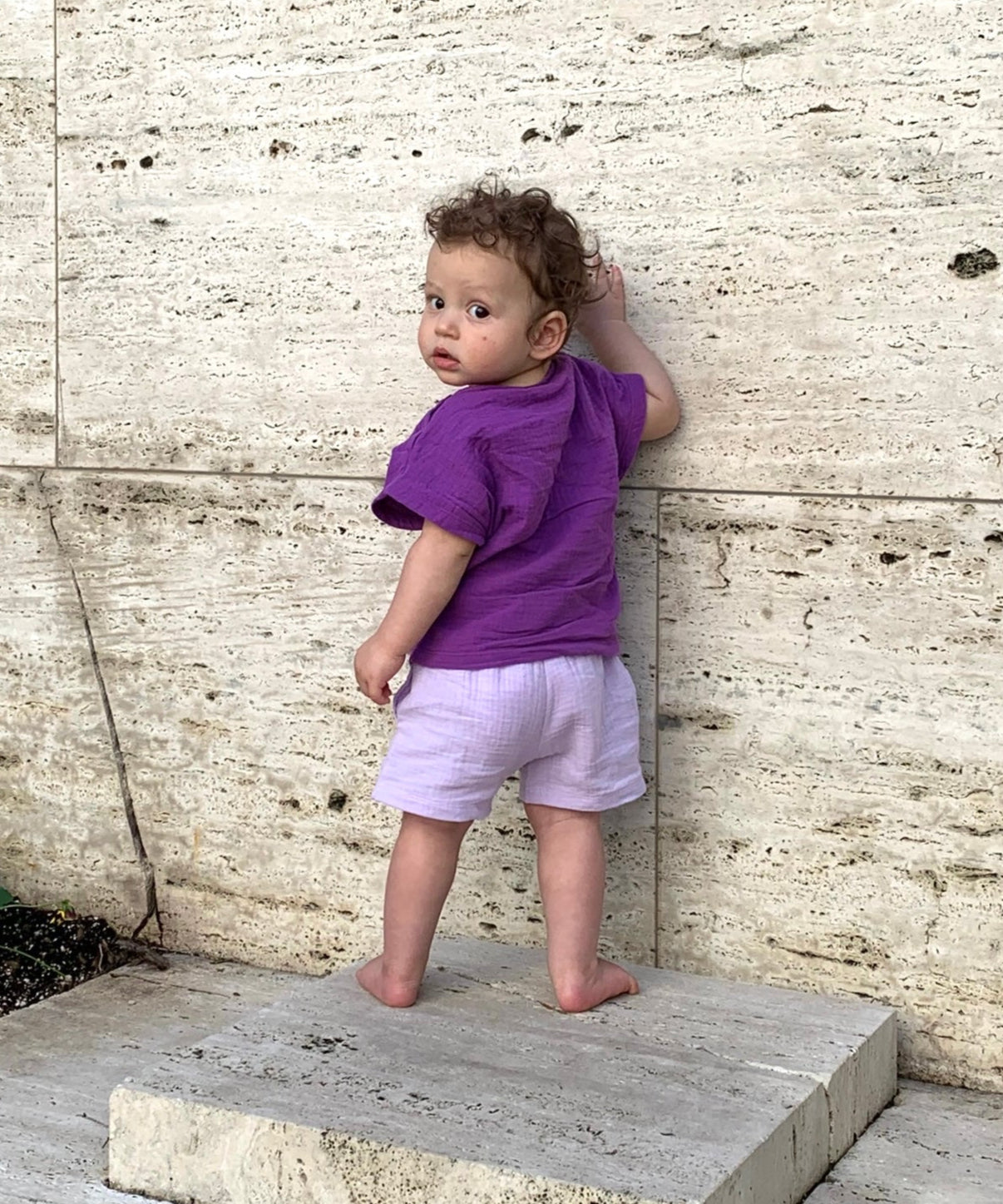 Child in a purple shirt and shorts standing on a stone step against a textured wall.