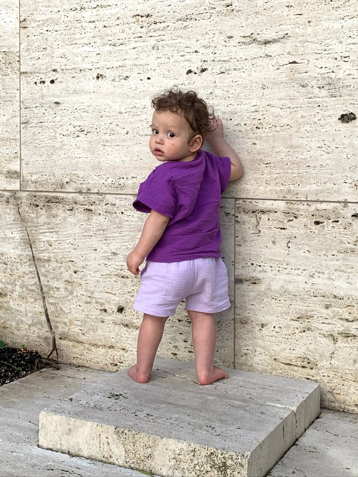 Child in a purple shirt and shorts standing on a stone step against a textured wall.