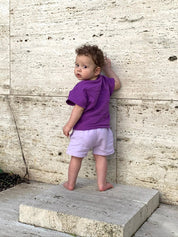 Child in a purple shirt and shorts standing on a stone step against a textured wall.